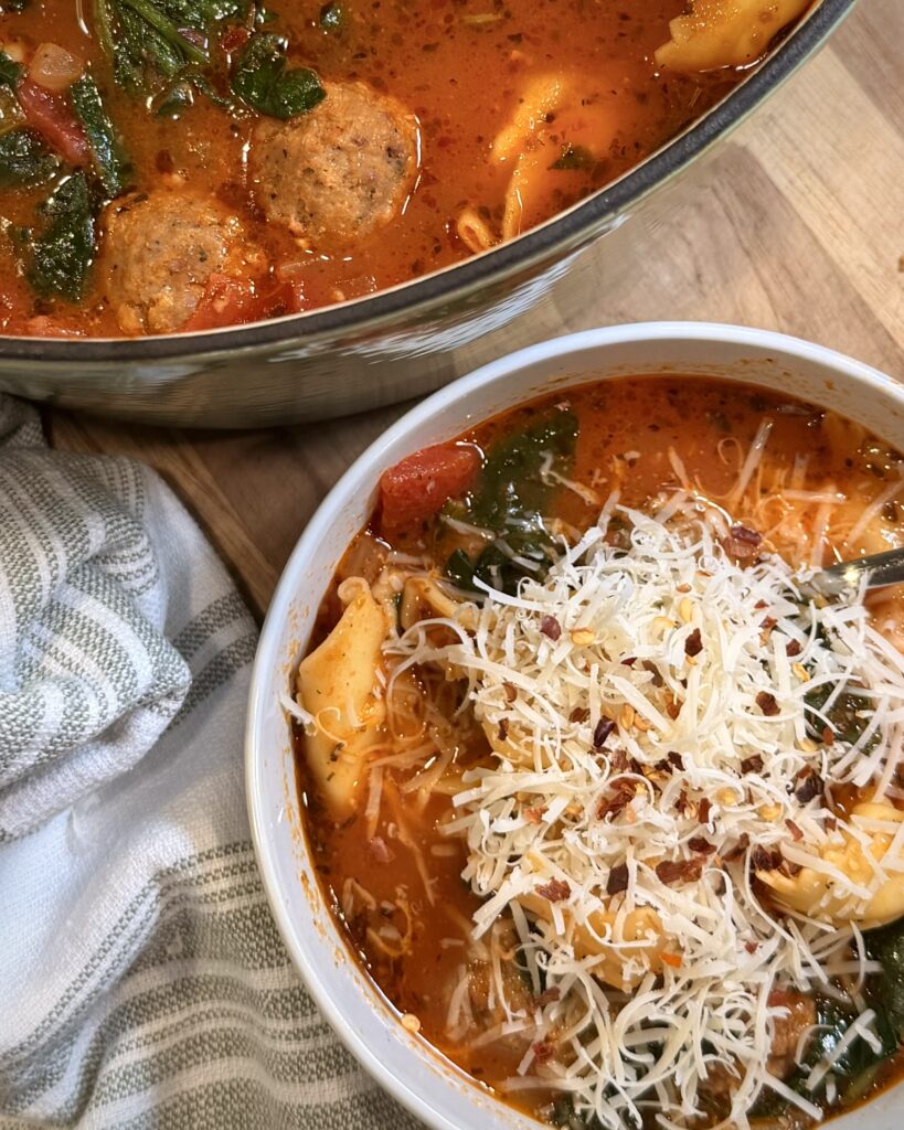 Tortellini Soup from Keep It Simple Y’all Everyday in a bowl with parmesan cheese on top. The bowl is sitting on a table next to the pot with more soup. 
