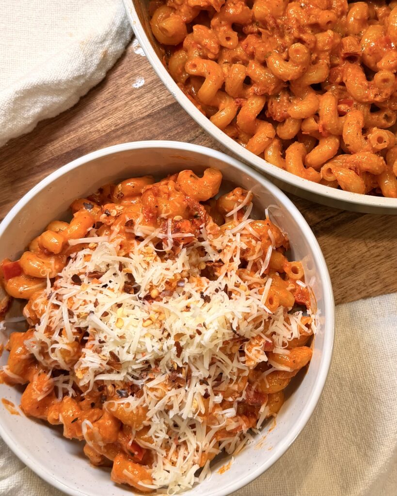 Creamy and rich Red Pepper Pasta sits in a bowl on a table next to a sliced red onion and sliced red bell pepper.