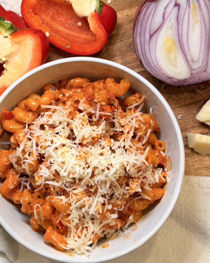 Creamy and rich Red Pepper Pasta sits in a bowl on a table next to a sliced red onion and sliced red bell pepper.