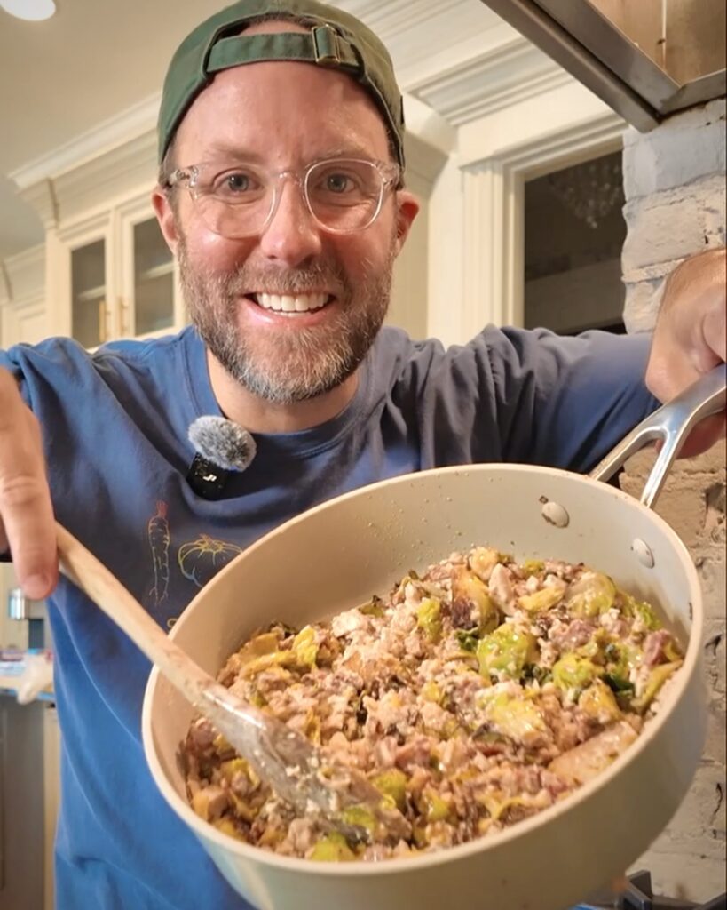 Matthew Bounds, creator of Your Barefoot Neighbor, holds a skillet with cooked Cheesy Bacon Chicken and Brussel Sprouts in his kitchen.