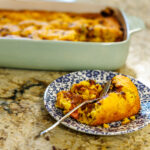 Your Barefoot Neighbor's easy Corndog Casserole sits on a plan on the counter next to the baking dish full of the hearty meal