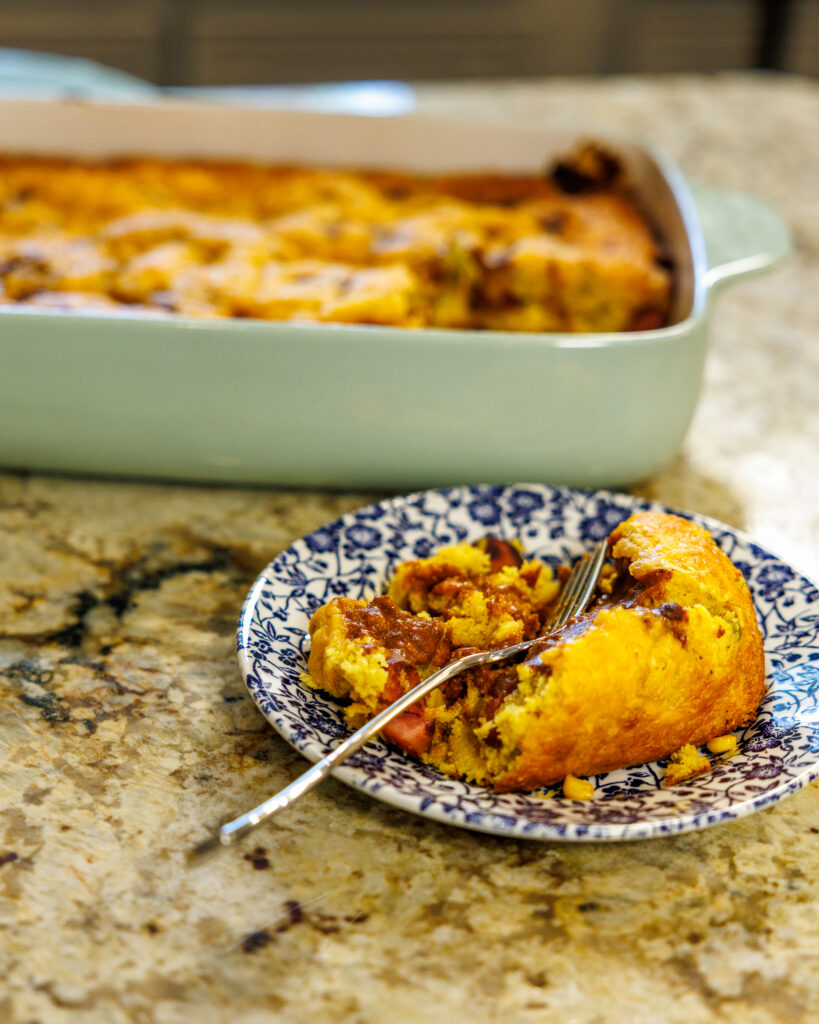 Your Barefoot Neighbor's easy Corndog Casserole sits on a plan on the counter next to the baking dish full of the hearty meal