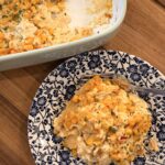 A generous helping of Your Barefoot Neighbor's creamy, cheesy Street Corn Enchilada Bake sits on a plate next to a baking dish with the casserole.