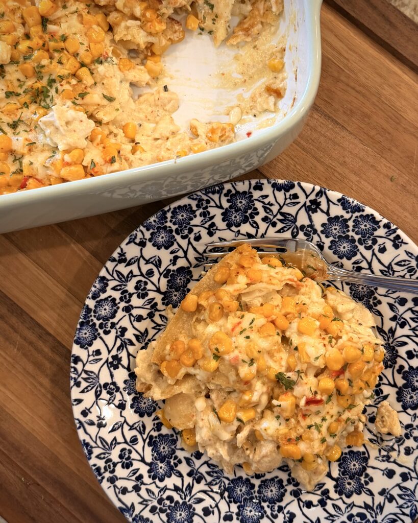 A generous helping of Your Barefoot Neighbor's creamy, cheesy Street Corn Enchilada Bake sits on a plate next to a baking dish with the casserole.