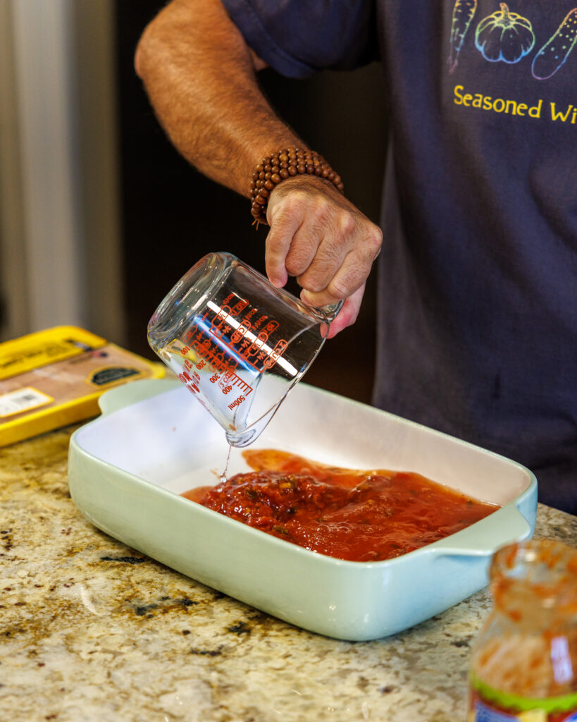 Your Barefoot Neighbor Matthew Bounds adds ingredients to the bottom of his pan for his Baked Swim Queso Chicken in his kitchen