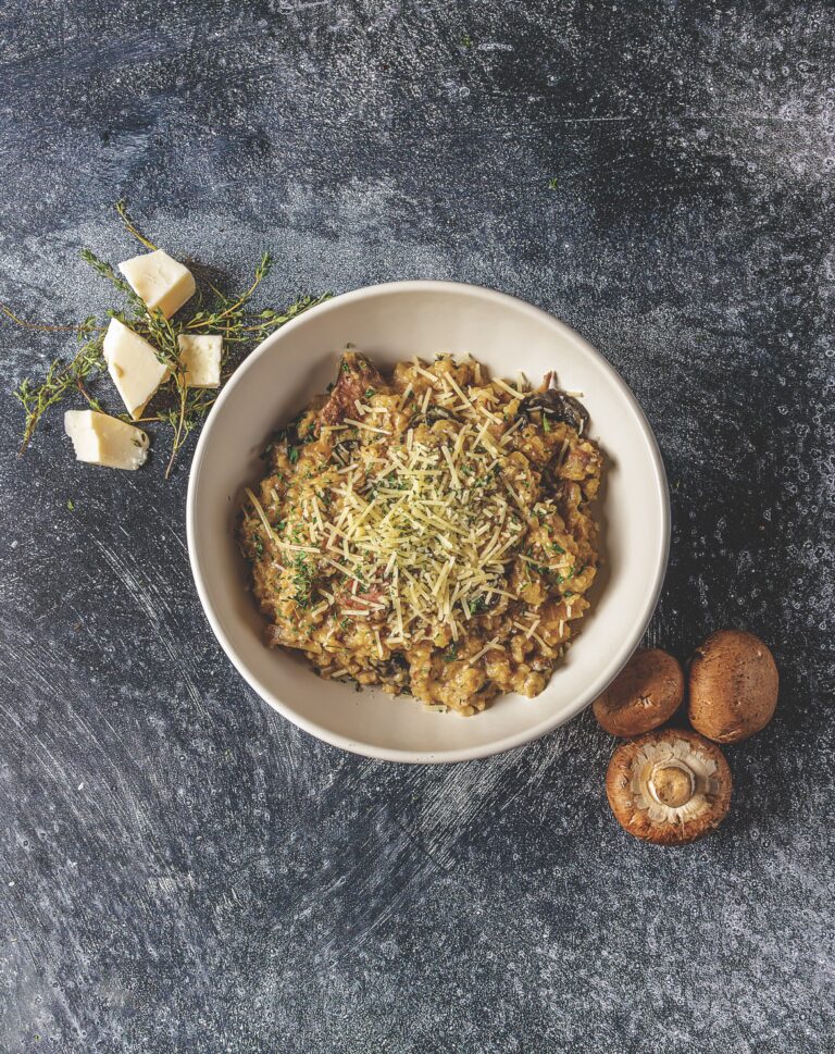 A generous helping of Your Barefoot Neighbor's Crockpot Beef and Mushroom Risotto is in a bowl on a dark stone countertop next to mushrooms