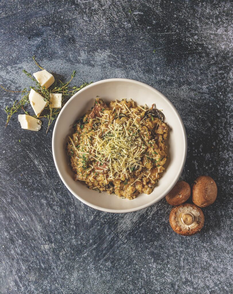 A generous helping of Your Barefoot Neighbor's Crockpot Beef and Mushroom Risotto is in a bowl on a dark stone countertop next to mushrooms