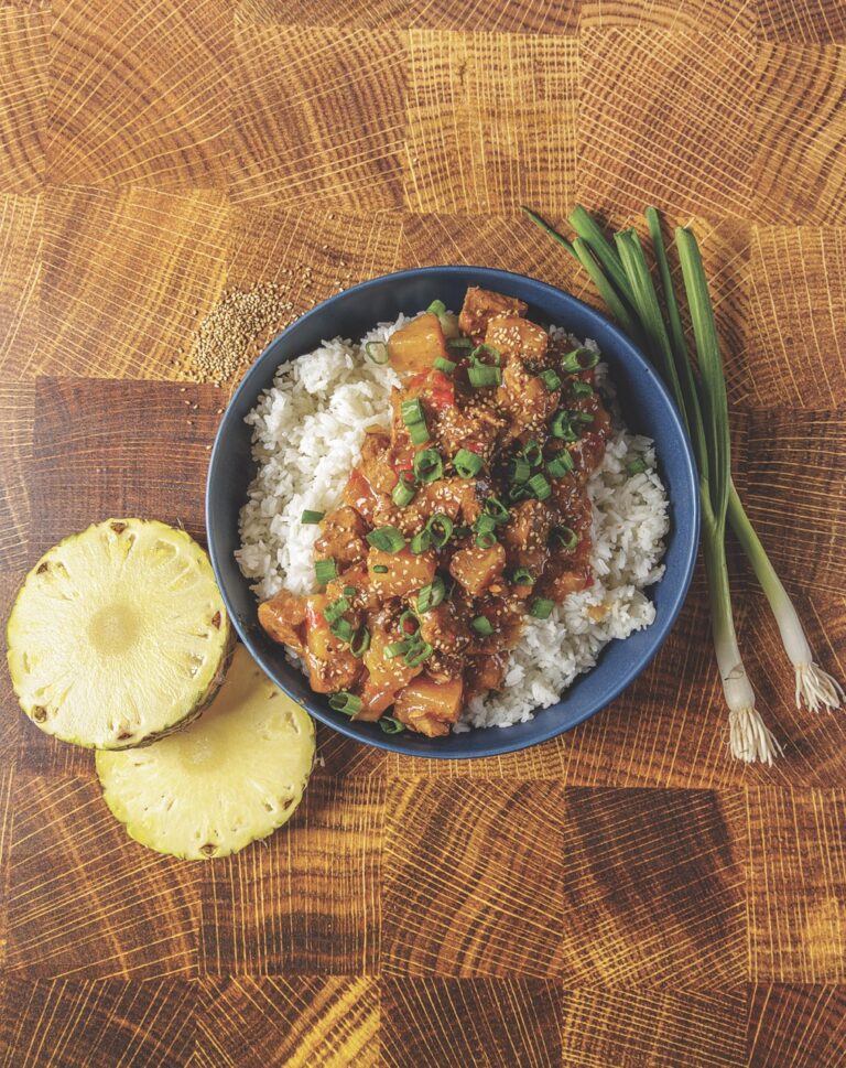 A large helping of Crockpot Hawaiian Pork made by Matthew Bounds, creator of Your Barefoot Neighbor, sits in a bowl on the counter next to green onions and pineapple slices