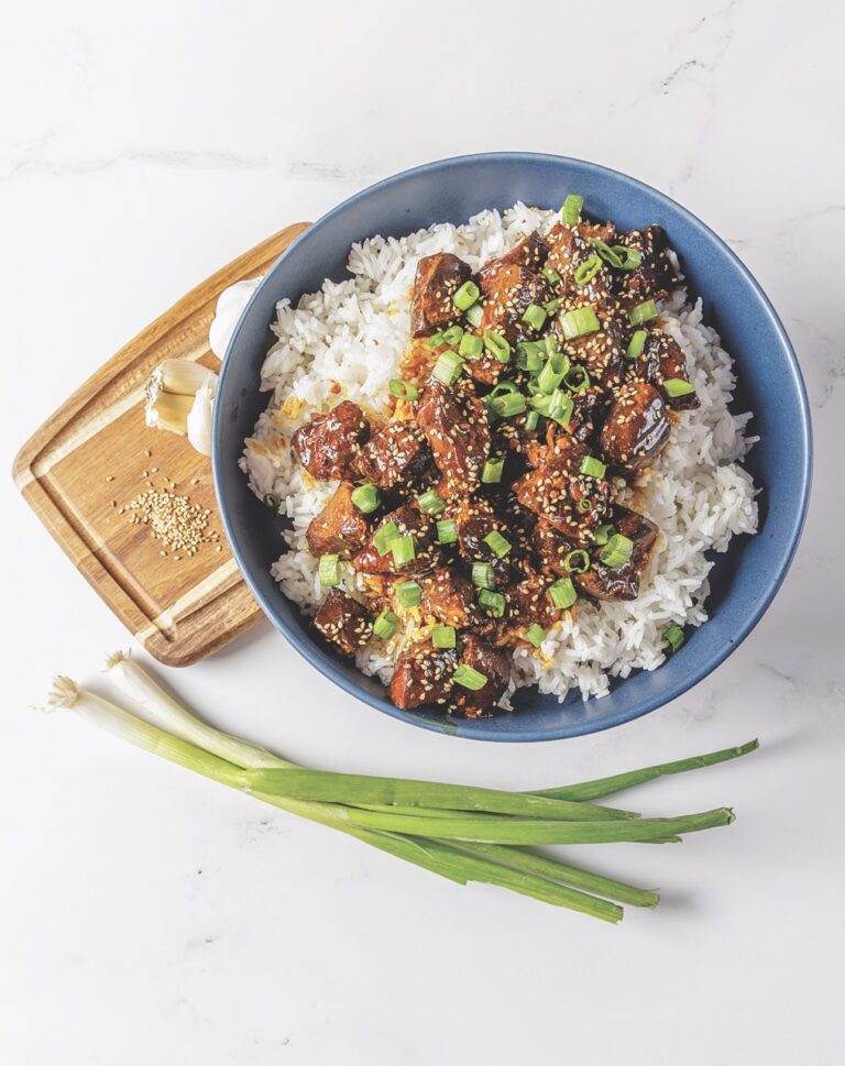A hearty helping of Your Barefoot Neighbor's crockpot Honey Garlic Chicken sits in a bowl with rice next to green onions.