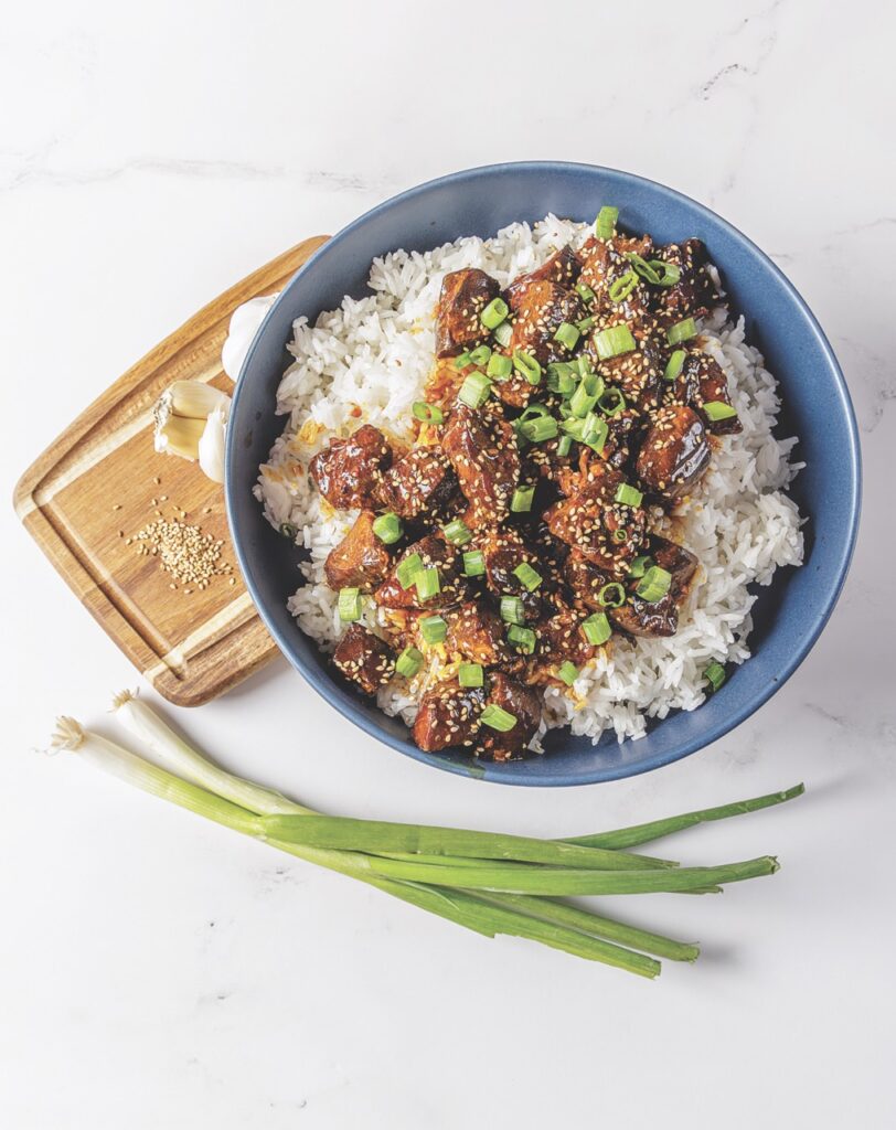 A hearty helping of Your Barefoot Neighbor's crockpot Honey Garlic Chicken sits in a bowl with rice next to green onions.