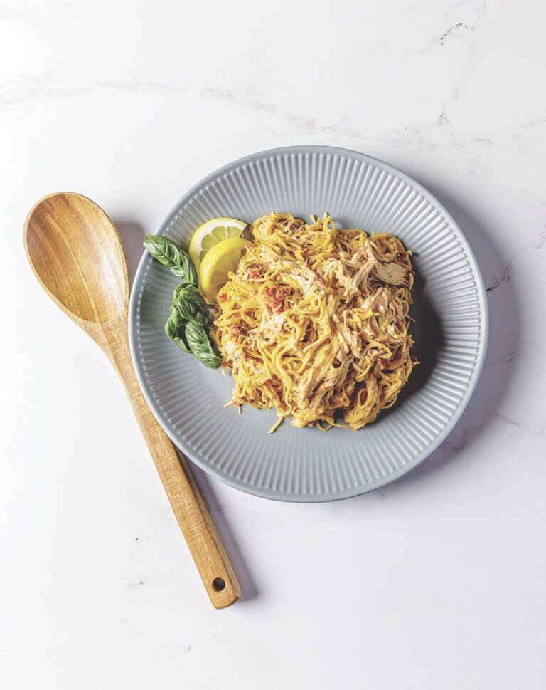 A large helping of Your Barefoot Neighbor Matthew Bounds' Sun Dried Lemon Tomato Chicken Pasta is on a plate on the counter next to a wooden serving spoon