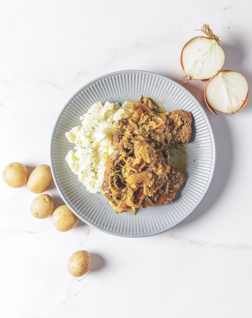 A generous helping of Your Barefoot Neighbor's Smothered Cube Steak and Onions is piled next to mashed potatoes on a plate next to fresh onions and potatoes.