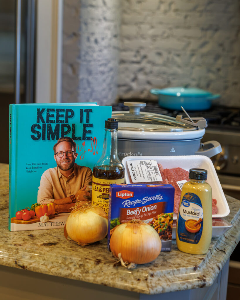 Ingredients for Your Barefoot Neighbor Matthew Bounds's Smothered Cube Steaks and Onions sit on the kitchen counter next to a Crockpot and Keep It Simple Y'all cookbook